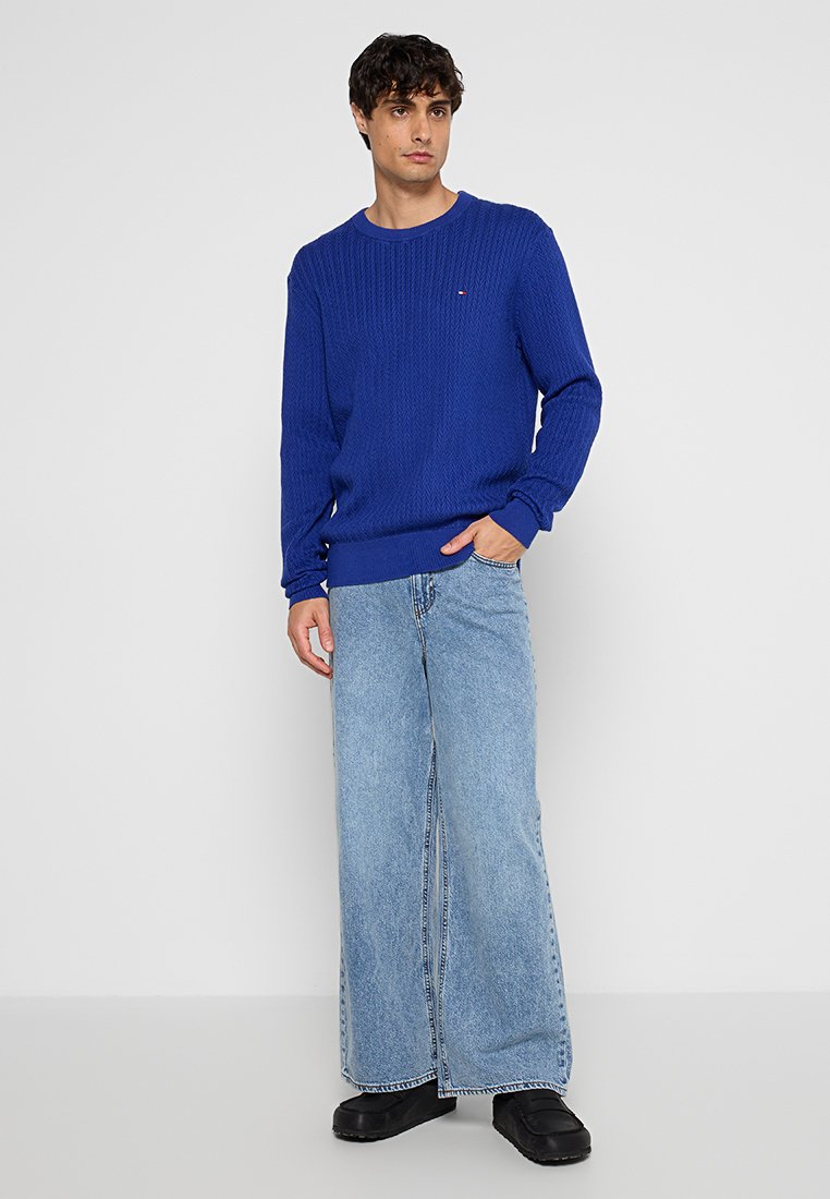 Blauwe gebreide trui met ribbelstructuur en ronde halslijn, gecombineerd met lichtblauwe denim jeans met wijde pijpen en zwarte schoenen.