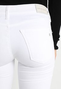 Jeans de denim blanco con un diseño ceñido. Las características incluyen un pequeño bolsillo trasero con un logotipo metálico y costuras blancas en contraste.