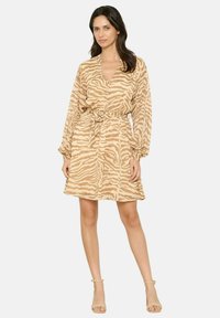 Frau in einem beigen und hellbraunen Zebrastreifen-Kleid mit langen Ärmeln, Taillengürtel und beigen Knöchelriemen-Pumps, die vor einem schlichten Hintergrund steht.