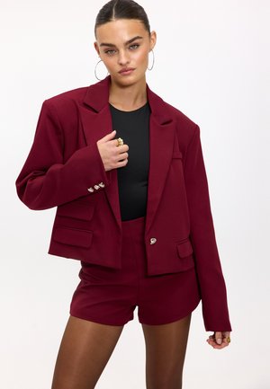 Bordeaux blazer met gestructureerde schouders, zilveren knoopaccenten en twee voorzakken, gecombineerd met bijpassende shorts in hetzelfde materiaal.