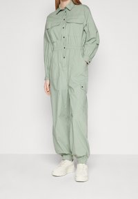 Mono verde claro con cintura ajustada, cierre de botones en el frente y dos bolsillos en el pecho. Puños y tobillos elásticos. Combinado con zapatillas blancas.