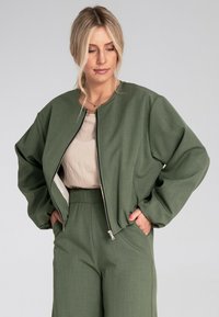 Zielona kurtka bombka z okrągłym dekoltem, z zapięciem na zamek i obszernymi rękawami, noszona na jasnobeżowej bluzce i pasujących spodniach.