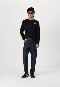 Black long-sleeve shirt with a yellow logo, paired with loose dark denim jeans and black sneakers with white stripes. Plain background.