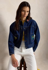 Femeie cu păr lung și închis la culoare, purtând o jachetă de blugi albastru închis cu petice brodate, o bluză galbenă și pantaloni albi, stând pe un scaun de lemn.