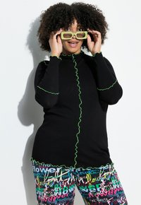 Zwarte top met lange mouwen en groene gerimpelde rand, gecombineerd met kleurrijke broek met patroon en tekst. Model draagt groene zonnebril.