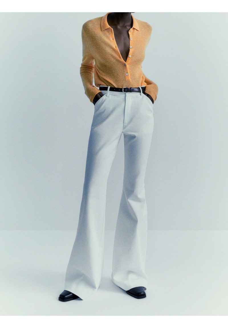 Personne portant un pull boutonné pêche rentré dans un pantalon large taille haute blanc avec une ceinture noire et des chaussures noires.