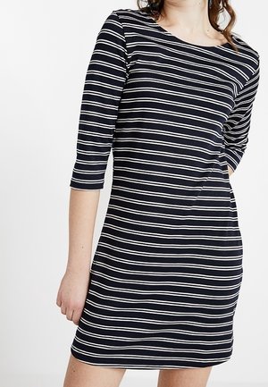 Kobieta w granatowej sukience z białymi poziomymi paskami i rękawami o długości do łokcia na jednolitym białym tle.