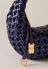 Navy-gewebte Handtasche aus glänzendem, strukturiertem Material, mit einem goldenen Hardware-Tag mit Logo und einem geflochtenen Henkel mit braunen Akzenten.