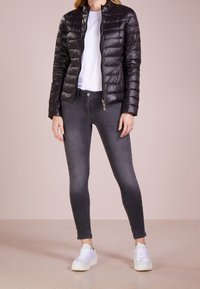 Sort pufferjakke med quiltet design, lynlås foran og høj krave, parret med tætsiddende mørkegrå jeans og hvide sneakers.