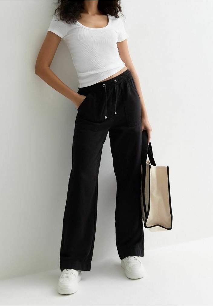 Zwarte wijde broek met elastische tailleband en trekkoord, gecombineerd met een strakke witte ribbeltop en witte sneakers. Canvas schoudertas inbegrepen.