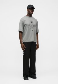 T-shirt en coton gris avec le texte "ULTRAS", associé à un pantalon ample noir avec un logo graphique. Porté avec des chaussures et une casquette noires.