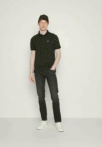 Ung mann iført svart polo skjorte med prikker, svarte jeans, hvite joggesko og svart caps, som står mot en enkel lys bakgrunn.