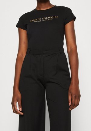 Persoon draagt een zwarte aansluitende t-shirt met de tekst "Armani Exchange Milano / New York" in goudkleur en een zwarte broek met hoge taille.