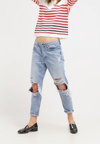 Jeans azul claro, rasgados con dobladillos deshilachados y grandes cortes en las rodillas, combinados con un suéter de rayas rojas y blancas y mocasines negros de charol.