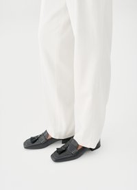 Μαύρα δερμάτινα loafers με τετράγωνη μύτη, λεπτομέρειες με φούντες και αντίθετες ραφές, συνδυασμένα με φαρδιά λευκά παντελόνια σε ανοιχτό φόντο.