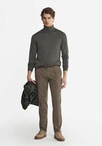 Grå polotröja i kombination med beige byxor, håller en mörkgrön jacka. Har en avslappnad passform och ljusbruna sneakers.