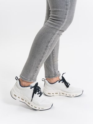 Person trägt hellgraue Skinny-Jeans und weiße Sportschuhe mit schwarzen Schnürsenkeln vor weißem Hintergrund.