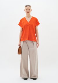 Top arancione a maniche corte con scollo a V e cucitura centrale, abbinato a pantaloni beige a gamba larga. Una piccola borsa marrone è tenuta in una mano.