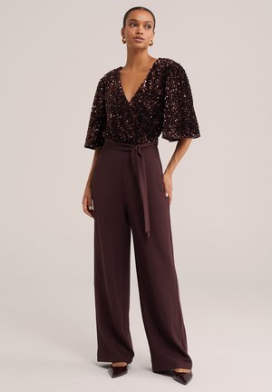 Femme portant un haut marron foncé à sequins avec des manches larges au niveau des coudes et un pantalon large assorti avec une ceinture à la taille et des talons foncés.