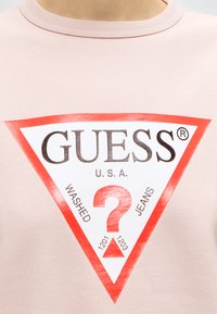 Chemise rose clair avec un logo triangulaire Guess rouge et blanc comportant un grand point d’interrogation et le texte « WASHED JEANS U.S.A. »