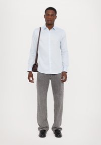 Cămașă albastră deschis cu nasturi, pantaloni gri texturați cu picioare largi și pantofi negri. Torbă maro tip crossbody, stând în fața unui fundal simplu.