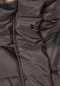 Chaqueta negra acolchada con una textura suave y brillante. Presenta secciones acolchadas y detalles ceñidos a lo largo de las costuras para un aspecto estructurado.