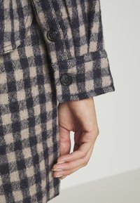 Mano descansando junto a la manga de un abrigo de lana con patrón de cuadros negro y beige y dos botones negros en el puño.