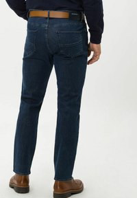 Jeans in denim blu scuro con vestibilità dritta, cinque tasche e dettagli di cucitura. Indossati con scarpe in pelle marrone e una cintura marrone.