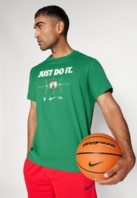 Žalias medvilninis marškinėliai su užrašu "JUST DO IT", krepšinio grafika ir NBA logotipu; dėvimi su raudonais sportiniais šortais, laikant oranžinį krepšinį.
