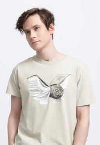 Lichtbeige katoenen t-shirt met een illustratie van een vliegende uil gemaakt van boekpagina's, met gedetailleerde veren en grote ogen.