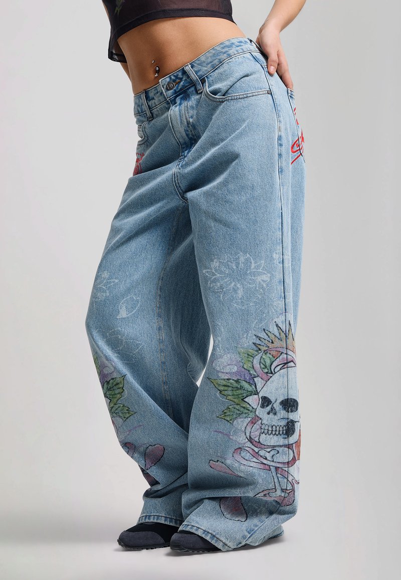 Ung person iført løse, lyseblå jeans med farverige kranie- og blomsterprint på benene, kombineret med sorte sneakers og en kort top.