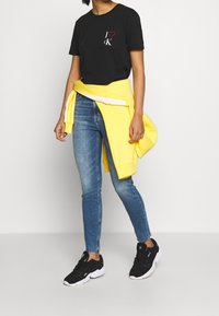 Czarna koszulka z napisem "I ❤️ ck", jasnoniebieskie obcisłe dżinsy, żółta bluza związana w talii i czarne sneakersy z białymi podeszwami.