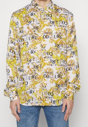 Chemise blanche à manches longues avec motifs baroques jaunes et texte noir portant l'inscription "Versace Jeans Couture", portée avec un jean bleu.