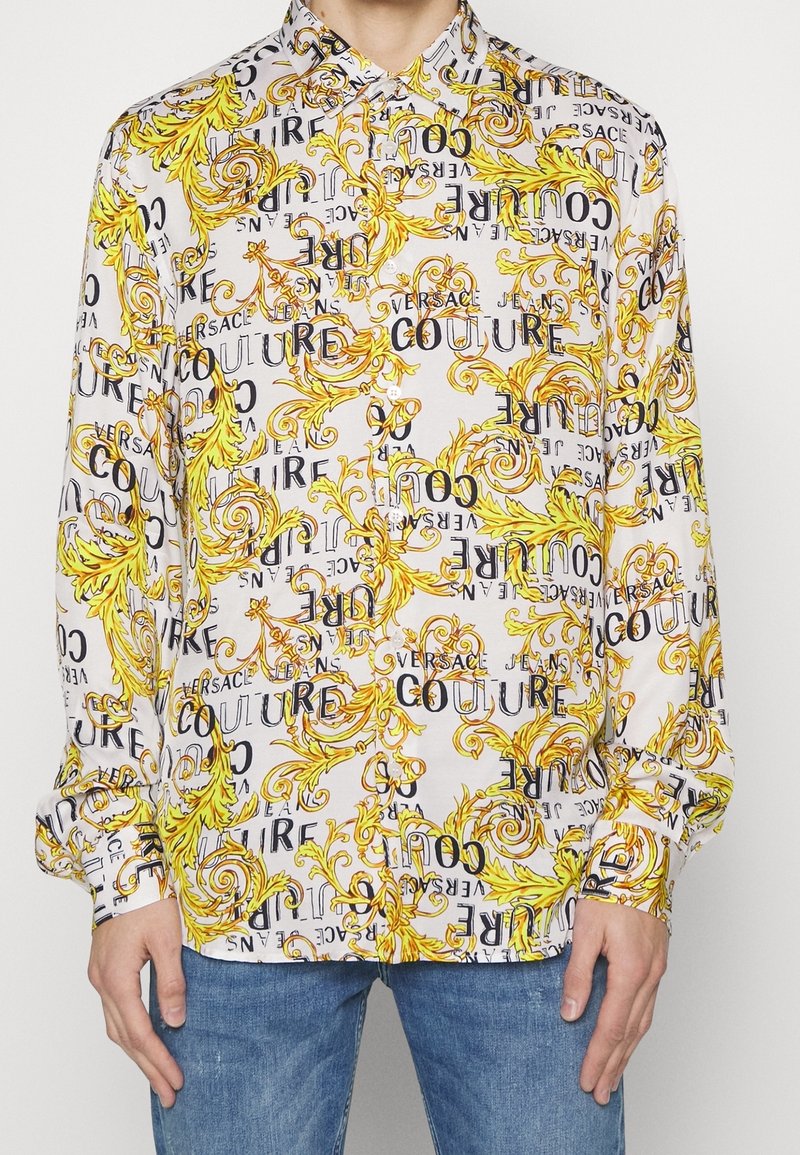 Chemise blanche à manches longues avec motifs baroques jaunes et texte noir portant l'inscription "Versace Jeans Couture", portée avec un jean bleu.