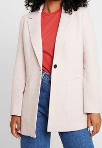 Jasnoróżowa marynarka o strukturze w jodełkę, z jednym czarnym guzikiem, klapami i dwoma bocznymi kieszeniami, nałożona na czerwoną koszulę.