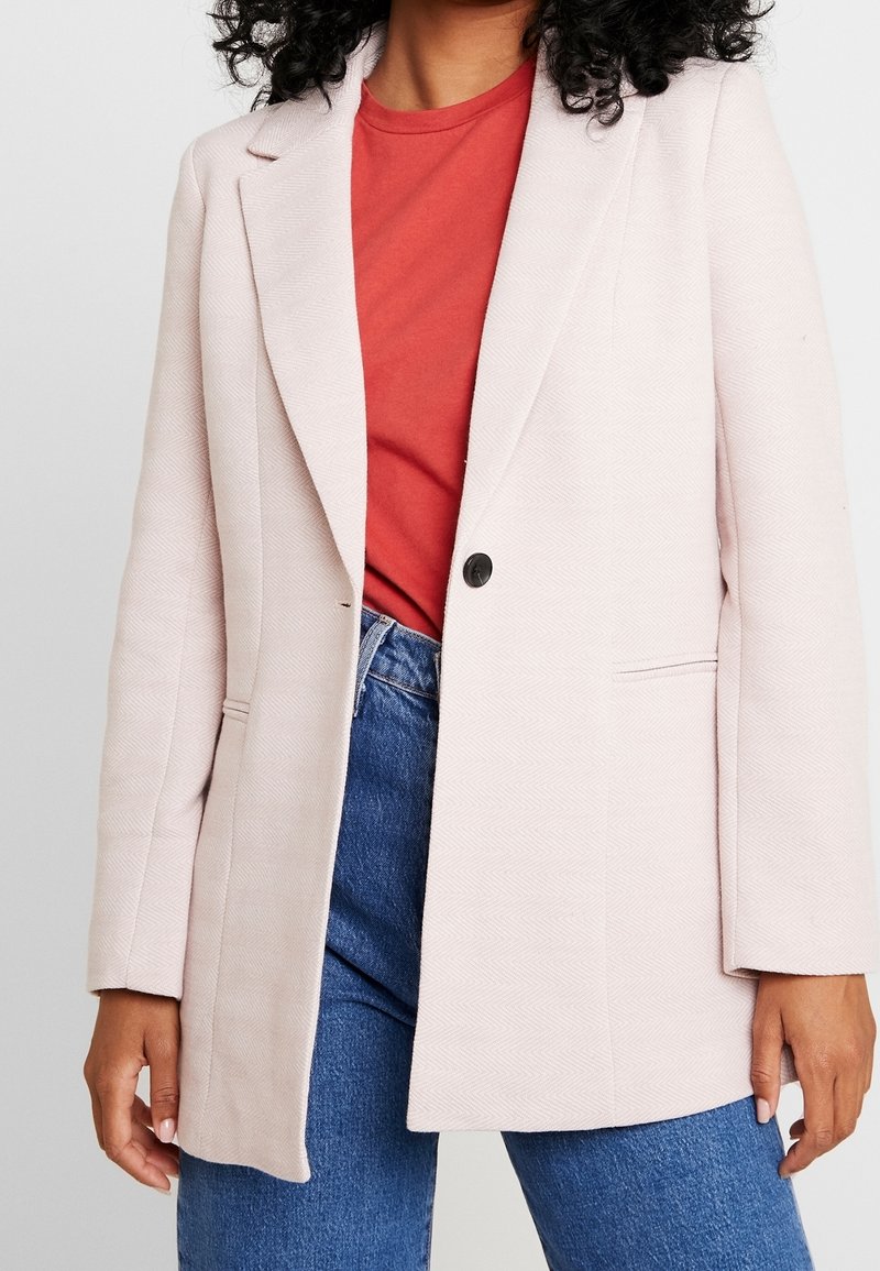 Jasnoróżowa marynarka o strukturze w jodełkę, z jednym czarnym guzikiem, klapami i dwoma bocznymi kieszeniami, nałożona na czerwoną koszulę.