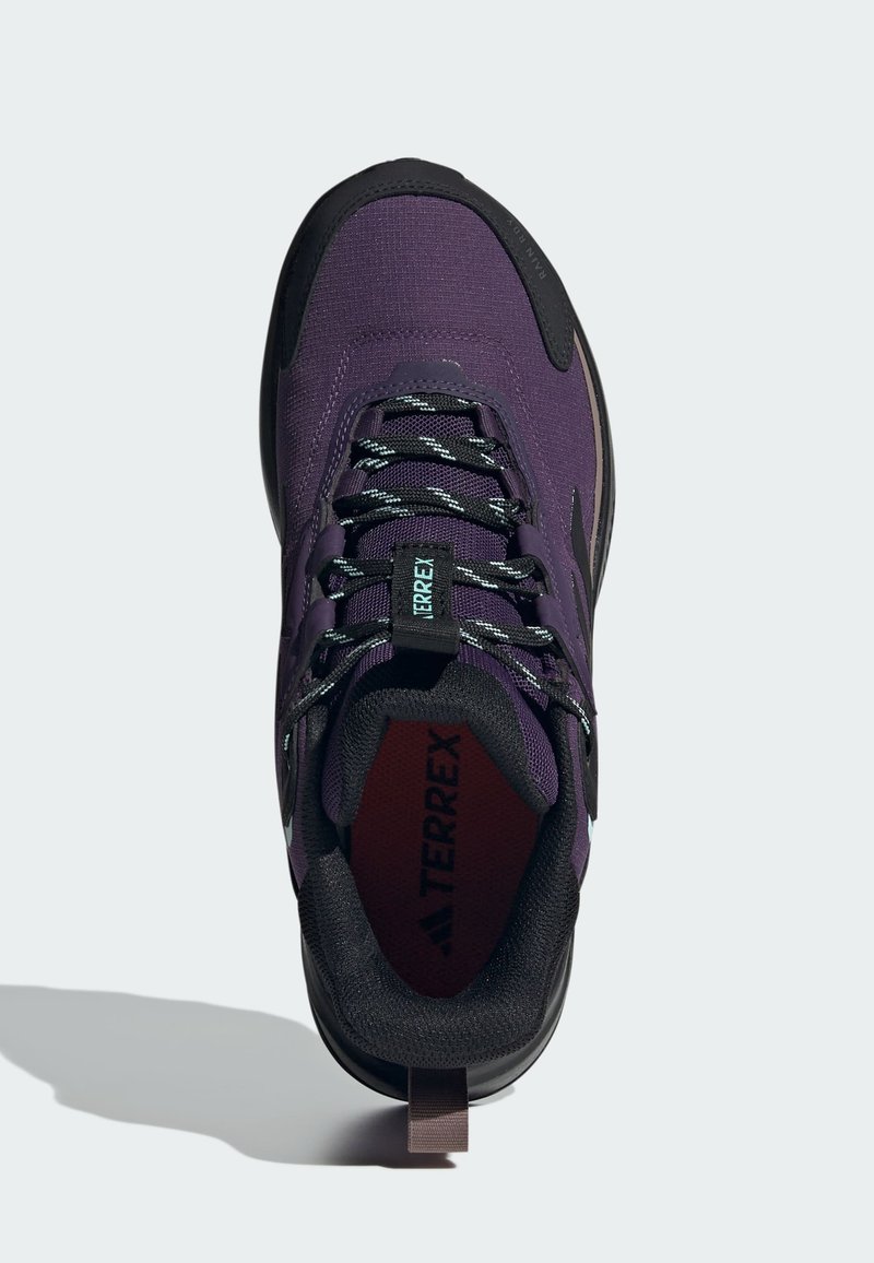 Zapatilla de senderismo morada con acentos negros, parte superior de tela texturizada, cordones reflectantes y un collar acolchado. Presenta una pestaña en el talón.