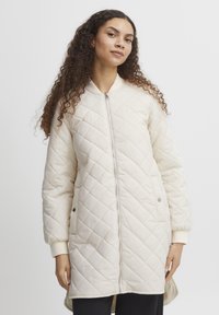 Quiltad krämfärgad lång jacka med dragkedja fram, ribbade muddarna och sidofickor. Har en mjuk struktur och diamantformat mönster.