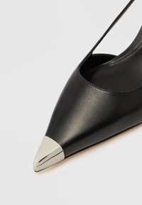 Tacón de cuero negro con punta afilada y detalle de tapa metálica plateada. Presenta un diseño recortado en los laterales para una apariencia elegante.