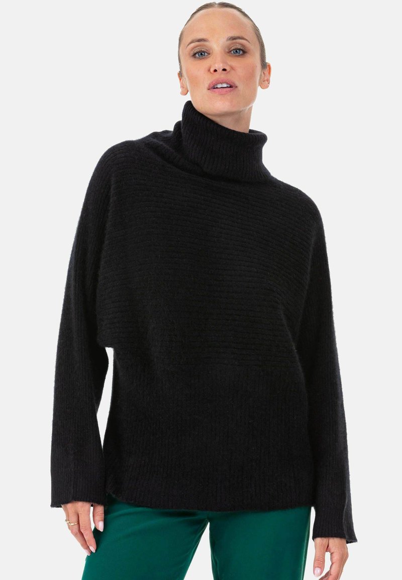 Pull noir oversize à col roulé avec un motif en maille texturée. Manches longues et coupe décontractée, porté avec un pantalon vert.