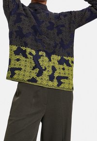 Persona con un maglione lavorato a maglia a motivo blu navy e verde oliva abbinato a pantaloni grigi scuri, braccia sollevate sopra la testa.
