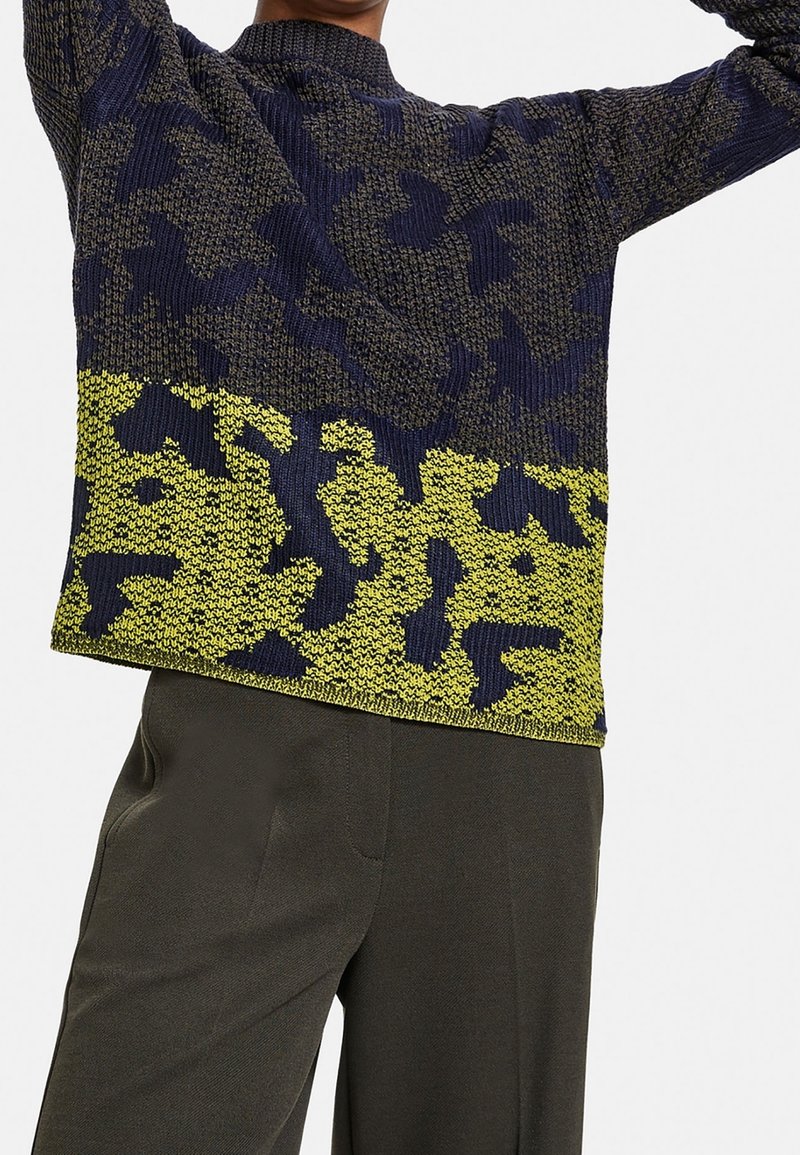 Persona con un maglione lavorato a maglia a motivo blu navy e verde oliva abbinato a pantaloni grigi scuri, braccia sollevate sopra la testa.