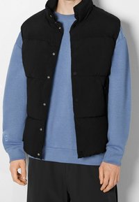 Černá péřová vesta s vysokým límcem, patentkami a prošívaným designem. Nosená přes světle modrou mikinu s hladkou texturou látky.