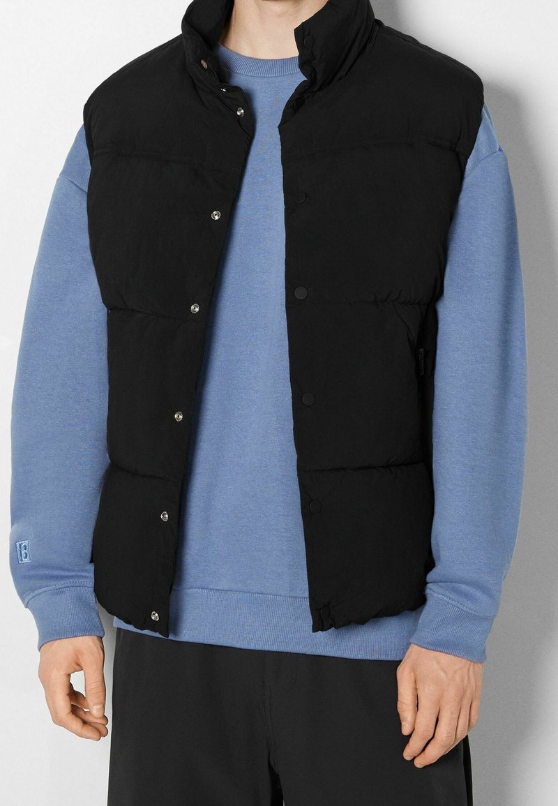 Černá péřová vesta s vysokým límcem, patentkami a prošívaným designem. Nosená přes světle modrou mikinu s hladkou texturou látky.