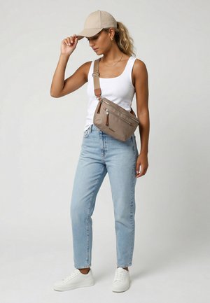 Kvinde iført en beige kasket, hvid tanktop, lyseblå jeans, hvide sneakers og en beige crossbody taske, stående mod en ensfarvet baggrund.