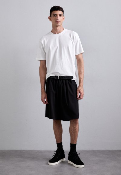 White short-sleeve t-shirt paired with a black pleated skirt. Black belt with silver buckle; complemented by black sneakers and ankle socks.