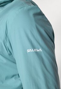 Ανοιχτό τιρκουάζ μπουφάν με λείο υφή, που διαθέτει το λευκό λογότυπο "SALEWA" στο αριστερό μανίκι και διακριτό κάθετο ριγωτό μοτίβο.