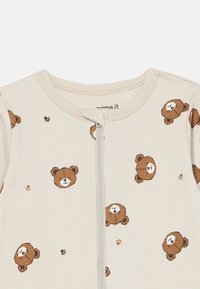 Lys kremfarget heldrakt med brune bjørn-trykk, med glidelås foran og mykt stoff. Rund halslinning uten ekstra detaljer.