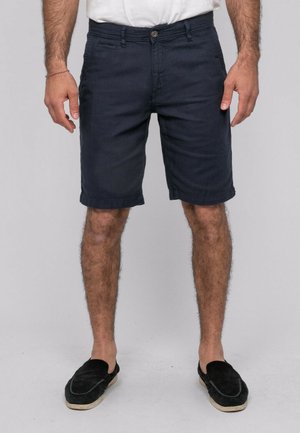 Homme portant un short bleu marine, un t-shirt blanc et des chaussures noires sans lacets, debout devant un fond uni.