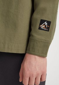 Zelená bavlněná rukáv s texturovaným povrchem, zdobený černou nášivkou s logem a oranžovým detailem. Ruka spočívá na tmavé látce.